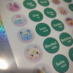 May include: A sticker sheet featuring circular designs with Animal Crossing character faces on light purple backgrounds and green circles with white text displaying names like Beau, Bob, Chai, Ione, Marshal, and Sasha alongside their specific ID numbers.