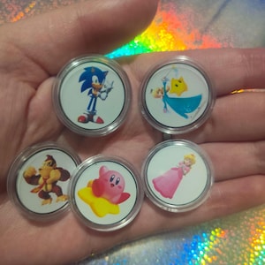 May include: Five circular plastic tokens featuring colorful character illustrations. The tokens display Sonic the Hedgehog, Rosalina with a yellow star, Donkey Kong, Kirby on a yellow star, and Princess Peach in a pink dress, all encased in clear plastic.