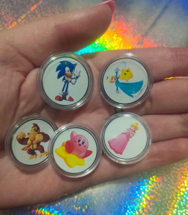 May include: Five circular plastic tokens featuring colorful character illustrations. The tokens display Sonic the Hedgehog, Rosalina with a yellow star, Donkey Kong, Kirby on a yellow star, and Princess Peach in a pink dress, all encased in clear plastic.
