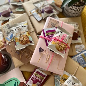 May include: A collection of wrapped gifts with a pink gift box in the centre that says "Mum" in gold wire letters. The gifts are wrapped in brown paper and decorated with twine, tea bags, and biscuits.