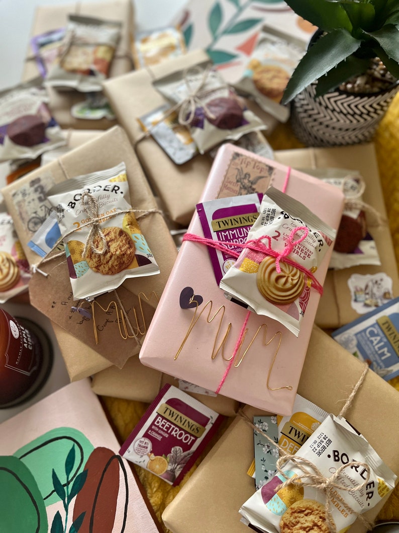 May include: A collection of wrapped gifts with a pink gift box in the centre that says "Mum" in gold wire letters. The gifts are wrapped in brown paper and decorated with twine, tea bags, and biscuits.