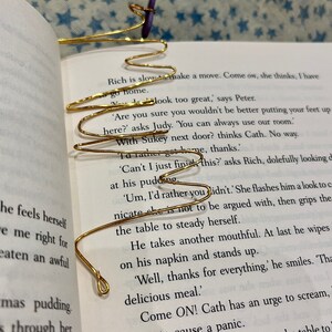 May include: A gold wire bookmark shaped like a wave, with a purple fabric bookmark attached to it. The bookmark is placed on an open book with text about a family dinner.
