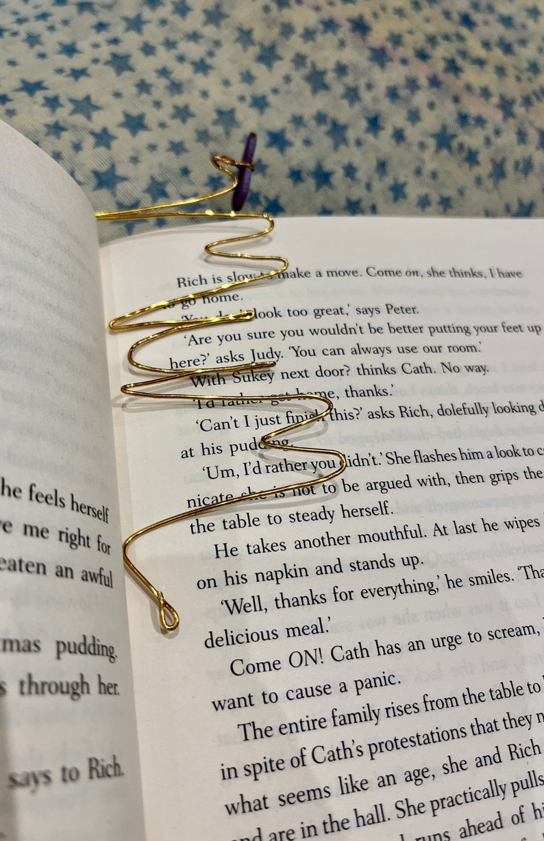 May include: A gold wire bookmark shaped like a wave, with a purple fabric bookmark attached to it. The bookmark is placed on an open book with text about a family dinner.