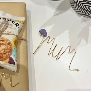 May include: A gold wire sculpture of the word "Mum" with a purple heart charm attached. The sculpture is on a white surface.