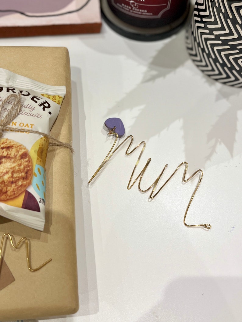 May include: A gold wire sculpture of the word "Mum" with a purple heart charm attached. The sculpture is on a white surface.