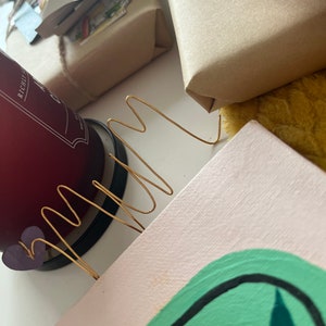 May include: A close-up of a pink canvas with a green abstract painting. A gold wire sculpture is placed on top of the canvas. The canvas is sitting on a table with a red candle and a brown package in the background.