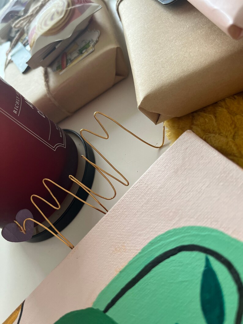 May include: A close-up of a pink canvas with a green abstract painting. A gold wire sculpture is placed on top of the canvas. The canvas is sitting on a table with a red candle and a brown package in the background.