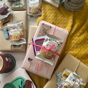 May include: A pink gift box wrapped with pink string and a gold wire that says "Mum". The box is surrounded by brown paper gift boxes, some with tea bags and biscuits inside. The boxes are on a yellow knitted blanket.