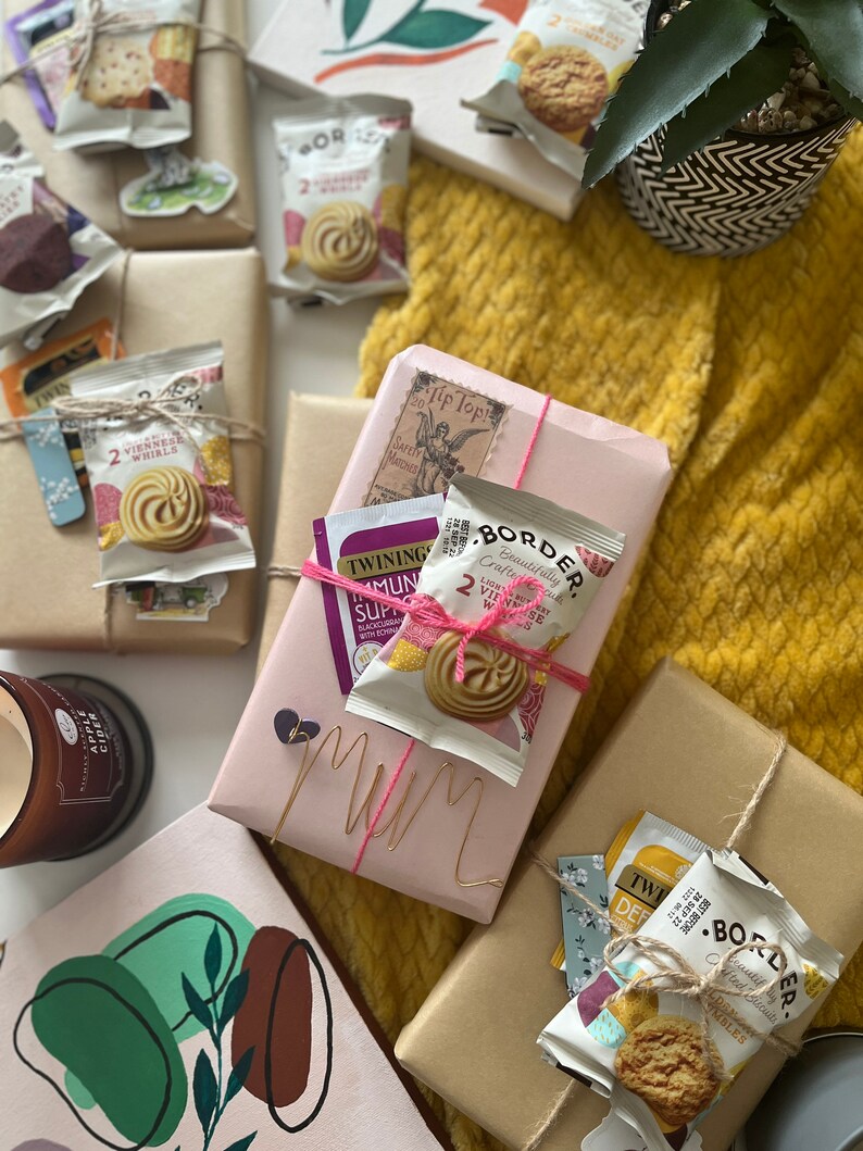 May include: A pink gift box wrapped with pink string and a gold wire that says "Mum". The box is surrounded by brown paper gift boxes, some with tea bags and biscuits inside. The boxes are on a yellow knitted blanket.