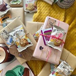 May include: A collection of wrapped gifts with brown paper, twine, and decorative tags. The gifts are arranged on a yellow knitted blanket. The gifts include tea, biscuits, and other treats. The gifts are labelled with the word "Mum".