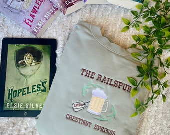 May include: A light green sweatshirt with the text "The Railspur Chestnut Springs" embroidered on the front. The sweatshirt also features a small embroidered patch with the text "Eaton" and a beer mug.