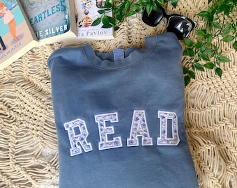 May include: A blue sweatshirt with the word "READ" embroidered on the front in white letters with a floral pattern. The sweatshirt is folded and lying on a white and beige crocheted blanket.