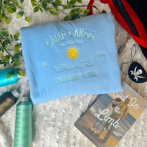 May include: A light blue sweatshirt with the text "Camp Cando" and "Swimming Camp" embroidered on the front. The sweatshirt also features a yellow sun and waves. The text "Est" and "2023" are also embroidered on the sweatshirt.