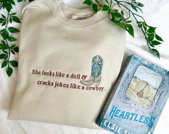 May include: A beige sweatshirt with embroidered text that reads "She looks like a doll & cracks jokes like a cowboy." The text is in brown and the image includes a brown cowboy boot with a blue design. The sweatshirt is laying on a white surface with a book titled "Heartless" by Elsie Silver.