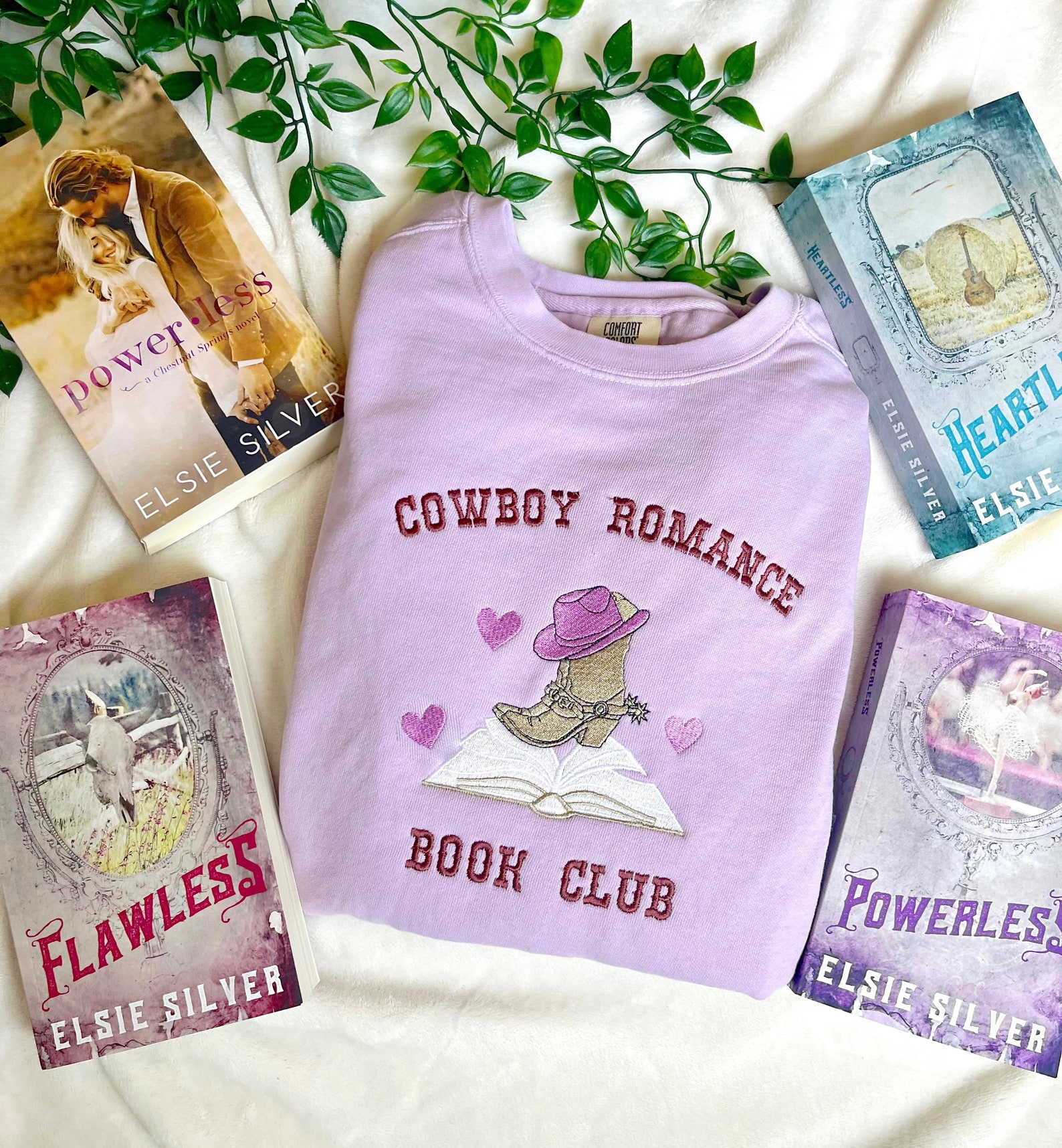 A folded purple sweatshirt with a crewneck. On the front of the sweatshirt is the text 'Cowboy Romance' in pink. An embroidered graphic of a cowboy boot and hat on top of an open book. Under the graphic is 'Book Club' in pink text. Multiple Elsie Silver titles surround the crewneck.