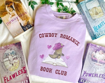 May include: A lavender sweatshirt with the text "Cowboy Romance Book Club" embroidered on the front. The embroidery includes a pink cowboy hat, boots, and a book. The sweatshirt is surrounded by four books with the titles "Powerless", "Heartless", "Flawless", and "Powerless" all by Elsie Silver.