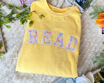 May include: A yellow sweatshirt with the word "READ" spelled out in floral fabric letters. The sweatshirt is surrounded by books and flowers.