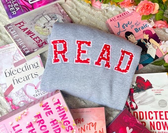 May include: A flat lay of a gray sweatshirt with the word "READ" in red letters with white polka dots. The sweatshirt is surrounded by pink and white books with titles like "Flawless", "Love on the Brain", "Things We Hide", and "The Infinity Between Us".