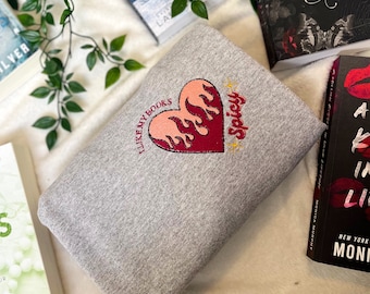 Könnte beinhalten: Ein graues Sweatshirt mit einem gestickten Herzdesign. Das Herz ist rot mit Flammenakzenten und dem Text "I like my books" und "Spicy". Das Sweatshirt ist gefaltet, mit Büchern im Hintergrund.