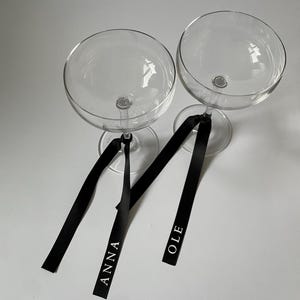 Könnte beinhalten: Zwei transparente Champagnergläser mit schwarzen Bändern. Auf den Bändern stehen die Namen "ANNA" und "OLE" in Weiß. Die Gläser haben eine runde Schale und einen schlanken Stiel.