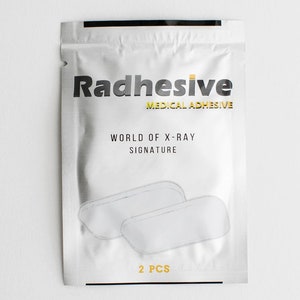 May include: A silver foil pouch with the text "Radhesive Medical Adhesive" and "World of X-Ray Signature" printed on it. The pouch contains two white, rectangular adhesive patches and the text "2 PCS" printed on the bottom.
