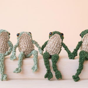 Könnte beinhalten: Vier handgefertigte Häkelfrösche in verschiedenen Grün- und Beigetönen. Jeder Frosch hat schwarze Augen und sitzt mit hängenden Beinen. Die Frösche halten sich an den Händen und bilden eine skurrile und charmante Darstellung.