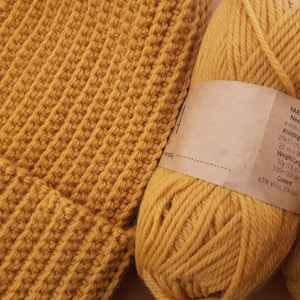 Könnte beinhalten: Zwei Knäuel senfgelber Wolle mit Etiketten, die die Wollgruppe, die Nadelstärke, die Strickspannung, das Gewicht/die Länge, den Inhalt und einen Prozentsatz an Wolle und Alpaka angeben. Die Wolle liegt neben einer gehäkelten senfgelben Mütze.