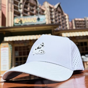 May include: White baseball cap with a black and white Mickey Mouse graphic on the front. The cap has a mesh back and a white brim.