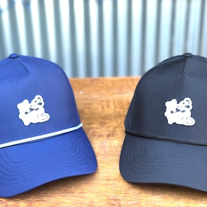 May include: Two baseball caps with a white logo of a bear wearing a bow tie. The cap on the left is blue and the cap on the right is black.