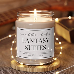 May include: A brown soy wax candle with a white label that reads "smells like FANTASY SUITES BACHELOR NATION APPROVED". The candle is lit and sitting on a wooden coaster.