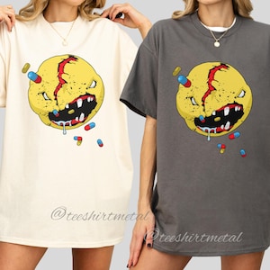 Op de afbeelding: Twee oversized T-shirts, één in naturel en één in houtskool, met een gele, boze gezichtsafbeelding met een rode scheur en witte tanden. Het ontwerp bevat verspreide pillen. De tekst "@teeshirtmetal" staat onderaan de shirts.