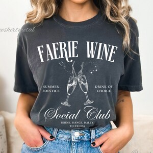 Könnte beinhalten: Ein dunkelgraues T-Shirt mit dem Text "FAERIE WINE" in weißen Buchstaben. Das Shirt zeigt außerdem eine Grafik von zwei Champagnergläsern, die aneinanderstoßen, mit dem Text "SUMMER SOLSTICE" und "DRINK OF CHOICE" über den Gläsern. Unter den Gläsern befindet sich der Text "Social Club" mit dem Text "DRINK. DANCE. DALLY. TO EXCESS" darunter.