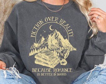 May include: A grey sweatshirt with a graphic print of a mountain range, a crescent moon, stars, and a stack of books. The text reads "Fiction Over Reality Because Romance Is Better In Books".