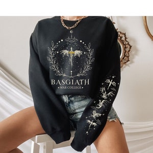 Puede incluir: Sudadera negra con un gráfico blanco de un dragón con las alas extendidas, rodeado de un círculo de estrellas y una corona. El texto "BASGIATH - WAR COLLEGE" está impreso debajo del gráfico.