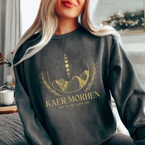 May include: A dark gray sweatshirt with a gold graphic design of mountains, a crescent moon, and stars. The text "Kaer Morhen Keep of the Elder Sea" is printed below the graphic.