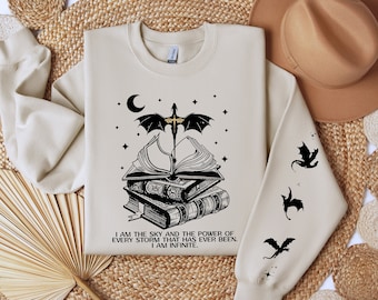 Può includere: Una felpa beige con una grafica nera di un drago che vola sopra una pila di libri. La grafica include una luna crescente e stelle. Il testo sulla felpa recita "Sono il cielo e il potere di ogni tempesta che sia mai esistita. Sono infinito."