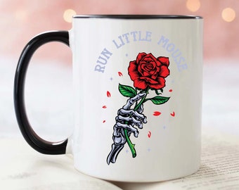 Puede incluir: Taza de café de cerámica blanca con borde y asa negros. La taza presenta un gráfico de una mano esquelética sosteniendo una rosa roja con el texto "Run Little Mouse" en un círculo morado.