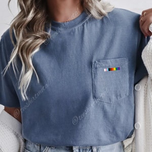 May include: A blue t-shirt with a pocket on the left side. The pocket has a rainbow stripe design.