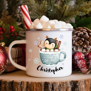 May include: White enamel mug with a penguin in a blue mug with marshmallows, cinnamon sticks, and a gingerbread cookie. The mug says "Merry Christmas" and "Christopher".