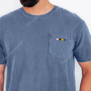 May include: A blue t-shirt with a pocket on the left side. The pocket has a small rainbow flag embroidered on it.