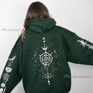 May include: A dark green hooded sweatshirt with a white graphic design on the back. The design features a circle with a crescent moon, a star, and a stylized eye. The sleeves have additional white graphic designs.