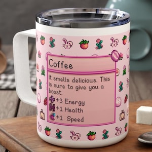 May include: White travel mug with a pink design featuring a game-like interface with the word "Coffee" and text. The mug has a silver rim and a white handle. The text says "It smells delicious. This is sure to give you a boost."