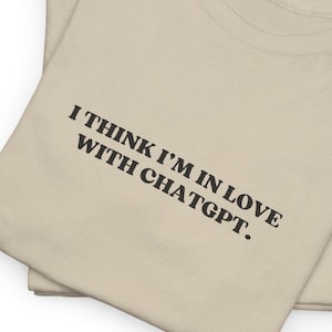 May include: A beige t-shirt with the text "I think I'm in love with ChatGPT." printed in black.