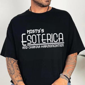 Peut inclure: T-shirt noir avec l'inscription blanche "Misty's Esoterica and Chakra Harmonization". Le t-shirt a des manches courtes et un col rond. Le th&egrave;me g&eacute;n&eacute;ral est spirituel et m&eacute;taphysique.