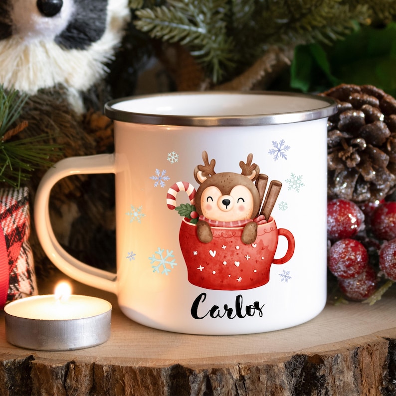 May include: White enamel mug with a red and white Christmas mug design featuring a cartoon reindeer inside the mug. The mug has a silver rim and handle. The text "Carlos" is printed on the mug.