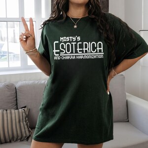 Peut inclure: T-shirt vert fonc&eacute; avec l'inscription blanche "Misty's Esoterica and Chakra Harmonization". Le t-shirt est de couleur unie et a une coupe ample. L'image montre le t-shirt port&eacute;.
