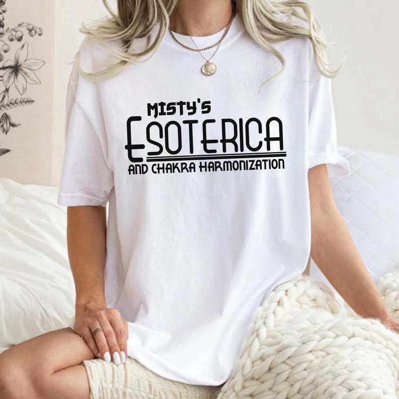 Peut inclure: T-shirt blanc avec l'inscription noire "Misty's Esoterica and Chakra Harmonization". Le t-shirt a des manches courtes et une coupe ample. Le texte est imprim&eacute; au centre sur le devant, offrant un design simple et &eacute;pur&eacute;.
