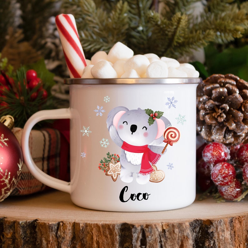 May include: White ceramic mug with a gray koala holding a red lollipop and wearing a red scarf. The mug has a snowflake pattern and the name "Coco" written on the bottom.