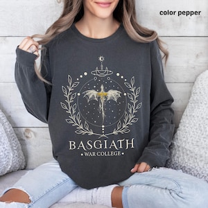 May include: A charcoal gray long-sleeved sweatshirt with a white graphic design of a dragon in a circle with stars and leaves. The text "BASGIATH WAR COLLEGE" is printed below the dragon.