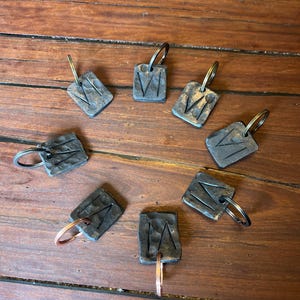 Puede incluir: Ocho llaveros de metal oscuro con una superficie texturizada y un diseño de la letra "V" están dispuestos en círculo sobre una superficie de madera. Cada llavero tiene un anillo de metal.